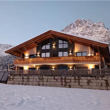 A beautiful chalet in the snow, surrounded by snow-covered mountains. The facade is made of wood and has large windows that allow for views outside.