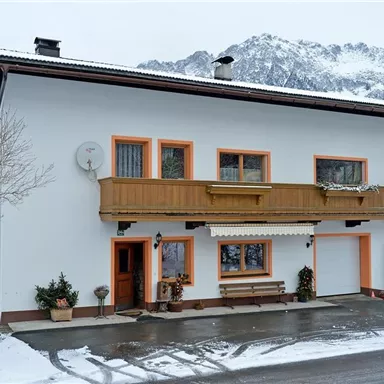 A cozy house in Alpine style with a wooden veranda. The snow covers the ground and the mountains in the background are visible.