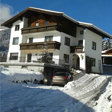 A modern house in the snow with wooden-clad balconies. The parking lot is covered with snow and the surroundings show wintery mountains.