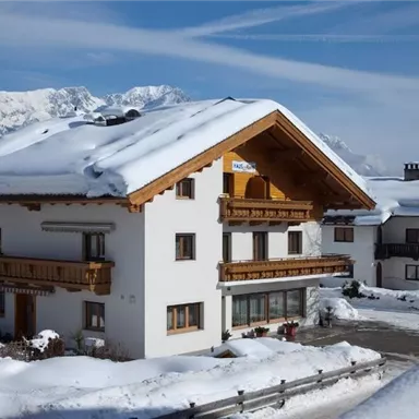 Ein charmantes Haus im alpinen Stil, umgeben von Schnee. Im Hintergrund sind schneebedeckte Berge und ein blauer Himmel zu sehen.