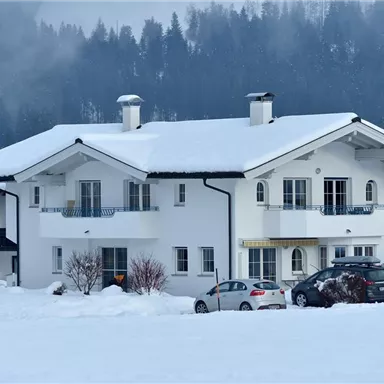 Ein schönes Haus im Schnee mit einem weißen Dach. Im Hintergrund sind verschneite Bäume und Berge zu sehen.
