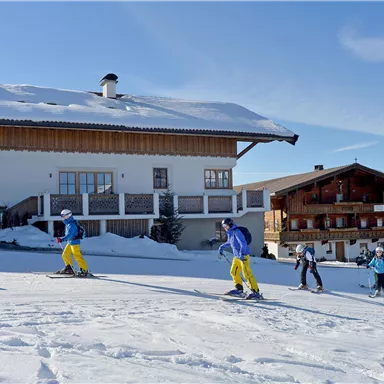 Eine Gruppe von Skifahrern geht in den Schnee, vor einem typischen Alpenhaus. Der Himmel ist klar und die Umgebung ist winterlich schön.