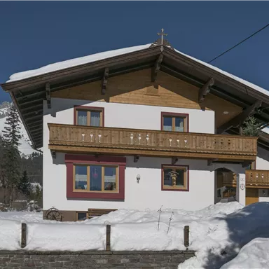 Ein charmantes Haus im Schnee mit einem Holzbalkon. Im Hintergrund sind schneebedeckte Berge und blauer Himmel zu sehen.