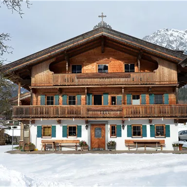 Ein traditionelles Holzhaus im bayerischen Stil, umgeben von Schnee. Im Hintergrund sind schneebedeckte Berge zu sehen.