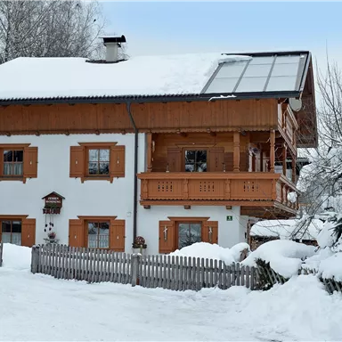 Ein charmantes Haus im alpinen Stil, umgeben von verschneitem Landschaft. Die Fensterläden und der Balkon aus Holz verleihen dem Gebäude einen gemütlichen Charakter.