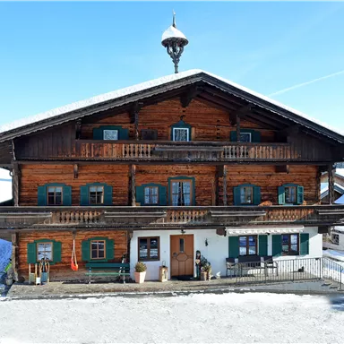 A traditional wooden house with several balconies and green windows. The surroundings are covered in snow and extend under a clear sky.