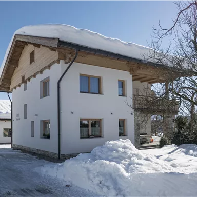 Ein modernes, zweigeschossiges Haus im Schnee mit einem Steildach und Holzverkleidung. Die Umgebung ist winterlich und ruhig, mit schneebedeckter Erde und einem klaren Himmel.