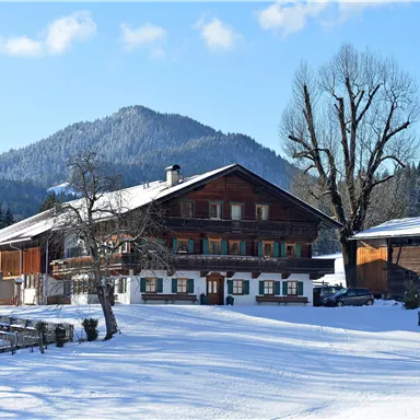 Ein traditionelles Holzhaus in einer verschneiten Landschaft. Im Hintergrund sind Berge und ein blauer Himmel zu sehen.