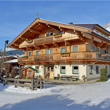 Ein charmantes, holzgebautes Haus im alpinen Stil, umgeben von Schnee. Im Hintergrund sind majestätische Berge und ein klarer blauer Himmel zu sehen.