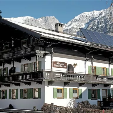 Ein traditionelles alpenländisches Haus mit einem schönen Balkon und grünen Fensterläden. Im Hintergrund sind schneebedeckte Berge zu sehen.