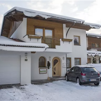 Ein modernes Haus im Schnee mit Holzverkleidung. Vor dem Haus stehen Autos und die Umgebung ist winterlich.