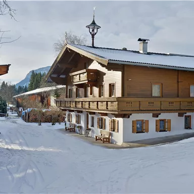 Ein malerisches Holzhaus im Winter mit Schnee bedecktem Boden. Im Hintergrund sind schneebedeckte Berge und ein klarer Himmel zu sehen.