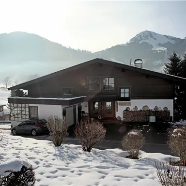 A charming house in the snow, surrounded by mountains and trees. The sun is shining, creating a warm atmosphere.
