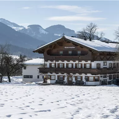 Ein traditionelles Haus im Alpenstil, umgeben von schneebedeckten Feldern. Im Hintergrund sind die Berge und ein klarer Himmel sichtbar.