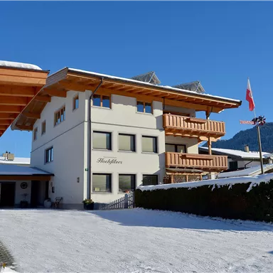 Ein modernes Gebäude im Schnee mit Holzverkleidung und Balkonen. Im Hintergrund sind Berge und ein blauer Himmel zu sehen.