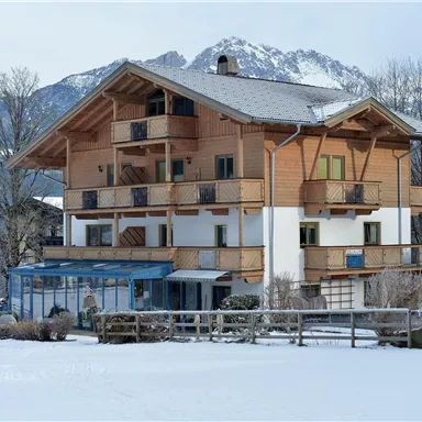 Ein schönes Holzhaus im Winter mit schneebedecktem Boden und den Alpen im Hintergrund. Die Bäume sind kahl und die Atmosphäre ist ruhig und malerisch.