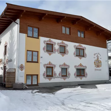 Ein traditionelles Haus mit einem Holzgeschoss und bunten Fenstern. Der Schnee liegt vor dem Gebäude, das von einem klaren Himmel umgeben ist.