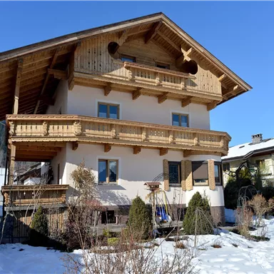 A charming wooden house with balconies and a snow-covered garden. In the background, mountains and a blue sky can be seen.
