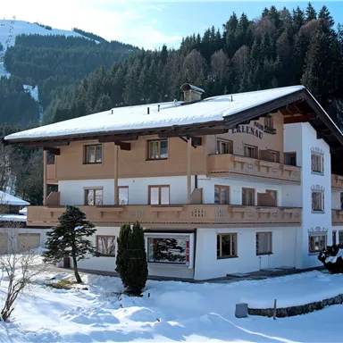 A modern building in the snow, surrounded by fir trees. Mountain peaks are visible in the background.