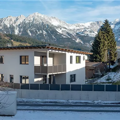 A modern house in the mountains, surrounded by snow-covered landscapes. In the background, impressive mountains lie under a clear sky.