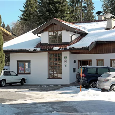 Ein weißes Haus mit einem roten Dach, umgeben von Schnee und Bäumen. Parkende Autos stehen vor dem Gebäude.