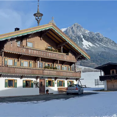 Ein traditionelles Holzhaus im Winter mit Schnee bedeckt. Im Hintergrund sind majestätische Berge und ein blauer Himmel zu sehen.