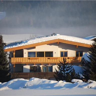 Ein gemütliches Haus mit einem Holzbalcon, umgeben von schneebedeckten Bäumen. Die Landschaft ist malerisch und winterlich.