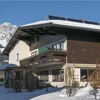 A cozy house in the snow, surrounded by impressive mountains. The clear blue sky complements the winter landscape.