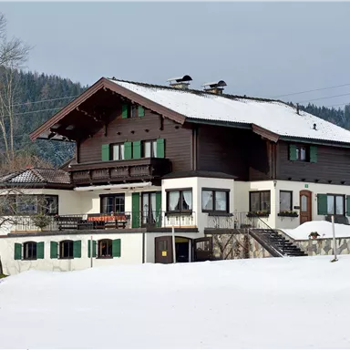 Ein großes, traditionelles Holzhaus im Schnee, umgeben von Bergen. Die Architektur weist typisch alpine Elemente auf.