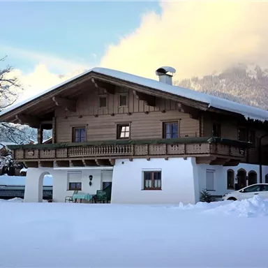 Ein traditionelles Holzhhaus im Schnee, umgeben von einer malerischen Winterlandschaft. Im Hintergrund sind verschneite Berge zu sehen.