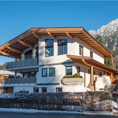 A modern house with wooden cladding and large windows, surrounded by snow-covered trees. In the background, mountains can be seen.