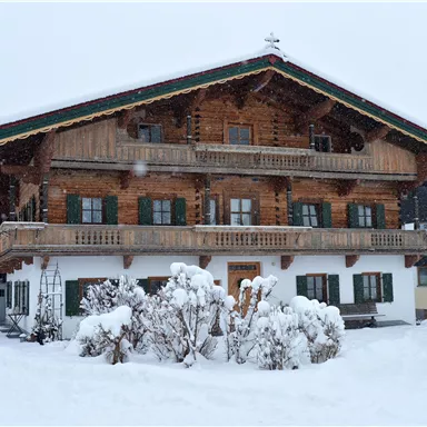 A traditional Alpine house with a wooden structure, surrounded by snowy terrain. The sky is gray and there are snowdrifts in front of the building.