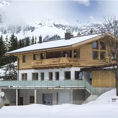 A modern wooden house in the mountains, surrounded by snow. The landscape is picturesque with tall firs and majestic mountains in the background.