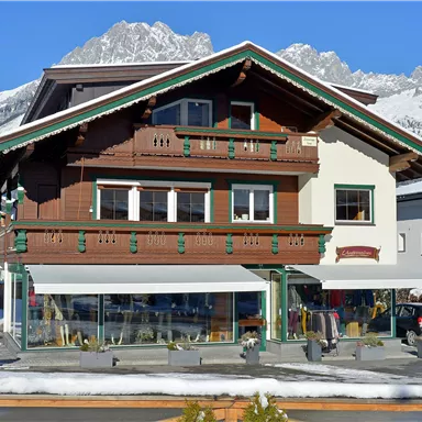 A traditional chalet with wooden cladding and balconies. In the background, snow-covered mountains and a clear blue sky are visible.