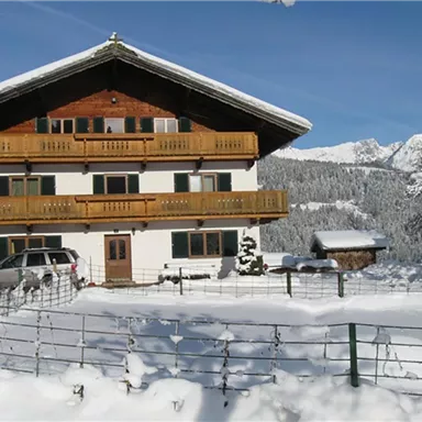 Ein gemütliches Haus im Schnee mit Holzbalkonen und einem klaren blauen Himmel. Die Landschaft ist winterlich und malerisch, umgeben von Bergen.
