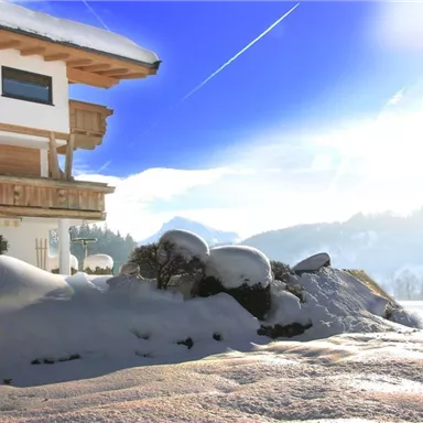 Ein gemütliches Haus in einer verschneiten Winterlandschaft. Der Himmel ist klar und blau, mit sanften Hügeln im Hintergrund.