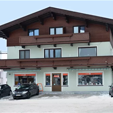 Ein zweigeschossiges Gebäude im alpinen Stil mit Holzverkleidung und Balkonen. Vor dem Haus parken mehrere Autos auf einem schneebedeckten Platz.