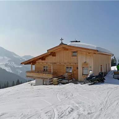 Eine gemütliche Hütte aus Holz steht auf einem schneebedeckten Hang. Im Hintergrund sind schneebedeckte Berge und eine Gruppe von Menschen zu sehen.