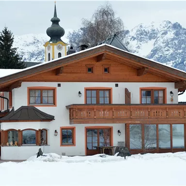 Ein traditionelles Haus im alpinen Stil, umgeben von Schnee. Im Hintergrund sind beeindruckende Berge zu sehen.
