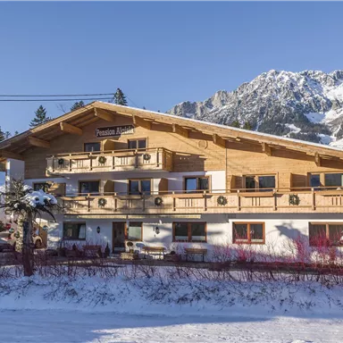 A cozy chalet in the snow, surrounded by mountains and trees. The clear blue sky complements the wintry landscape.