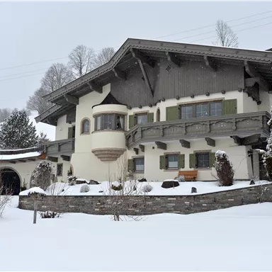 A charming alpine house in the snow, surrounded by snow-covered mountains. The architecture is traditional with a charming bay window and wooden embellishments.