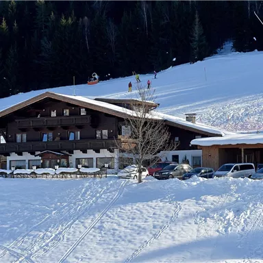 Ein Chalet inmitten einer schneebedeckten Landschaft. Im Hintergrund sind Skifahrer auf der Piste zu sehen.
