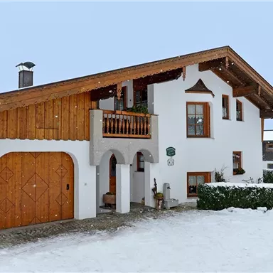 Ein charmantes Haus im alpinen Stil, umgeben von Schnee. Der Dachstuhl ist aus Holz, und es gibt eine kleine Terrasse mit Pflanzen.
