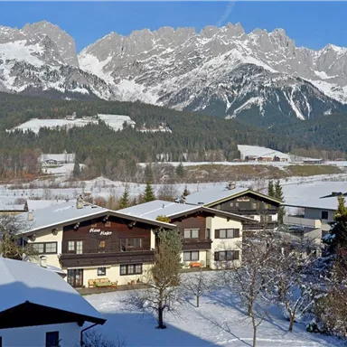 A picturesque winter landscape with snow-covered houses and majestic mountains in the background. The sky is clear and blue, emphasizing the serene atmosphere.