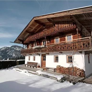 A traditional wooden house in the snow, surrounded by mountains. The architecture showcases a typical alpine style with a balcony and a well-maintained garden.