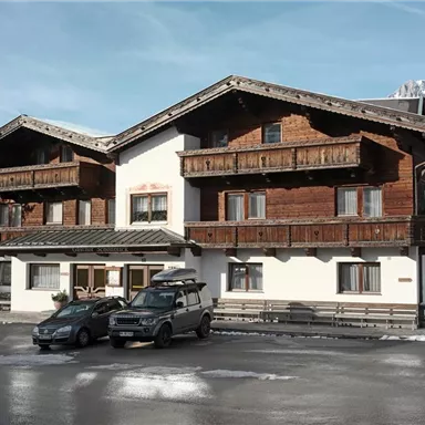 A typical alpine house with a wooden facade and balconies. In front of the house are several cars, surrounded by snow-covered ground.