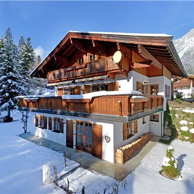 A beautiful chalet in the snow, surrounded by fir trees. In the background, mountains can be seen, the sky is clear and blue.