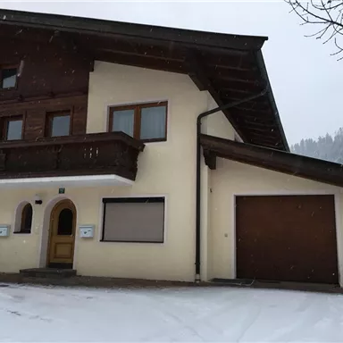 Ein traditionelles alpines Haus mit einem Holzbalcon und einem Garagentor. Schnee bedeckt den Boden und umgibt das Gebäude.