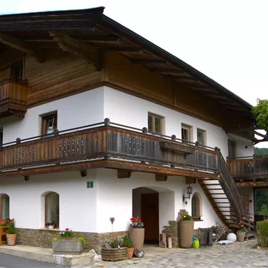 A traditional house in Alpine style with a wooden balcony. In the background, green mountains and a clear sky can be seen.