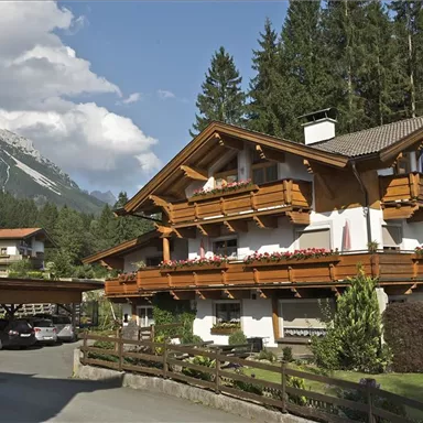 A traditional chalet surrounded by trees and mountains. The balconies are adorned with flowers, and the surroundings are peaceful and picturesque.
